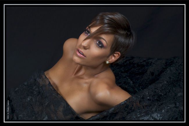 Modèle - Photographie portrait d'un modèle féminin, dont les contrastes vestimentaires et d'ambiance, sur différentes textures aux teintes noires, font ressortir le haut de son corps et son visage aux teintes bronzées